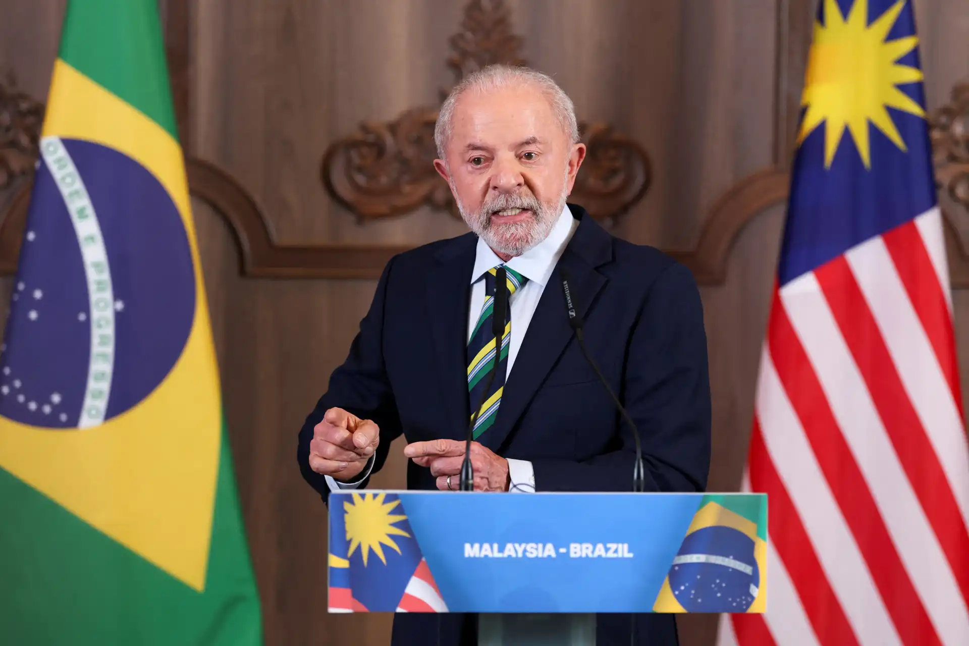 Elementos visuais de país, com bandeira brasileira e malásia, durante evento com discurso, destacando relações internacionais.