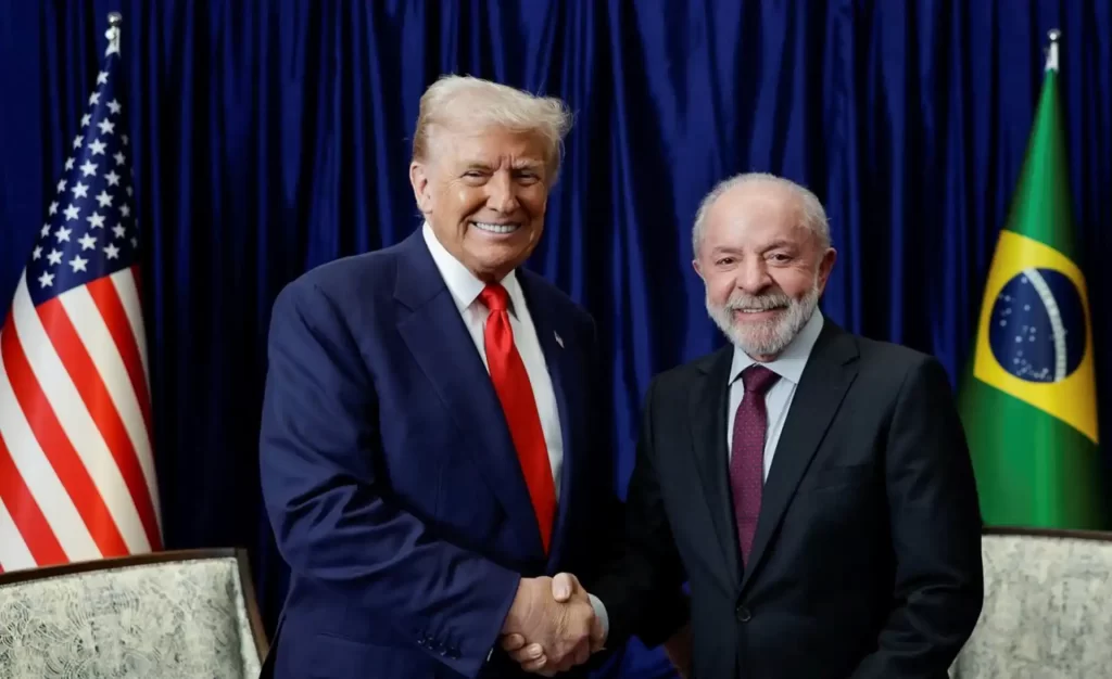 Imagem de Donald Trump e Luiz Inácio Lula da Silva sorrindo e apertando as mãos durante encontro oficial, com bandeiras dos Estados Unidos e Brasil ao fundo, simbolizando uma reunião entre líderes mundiais