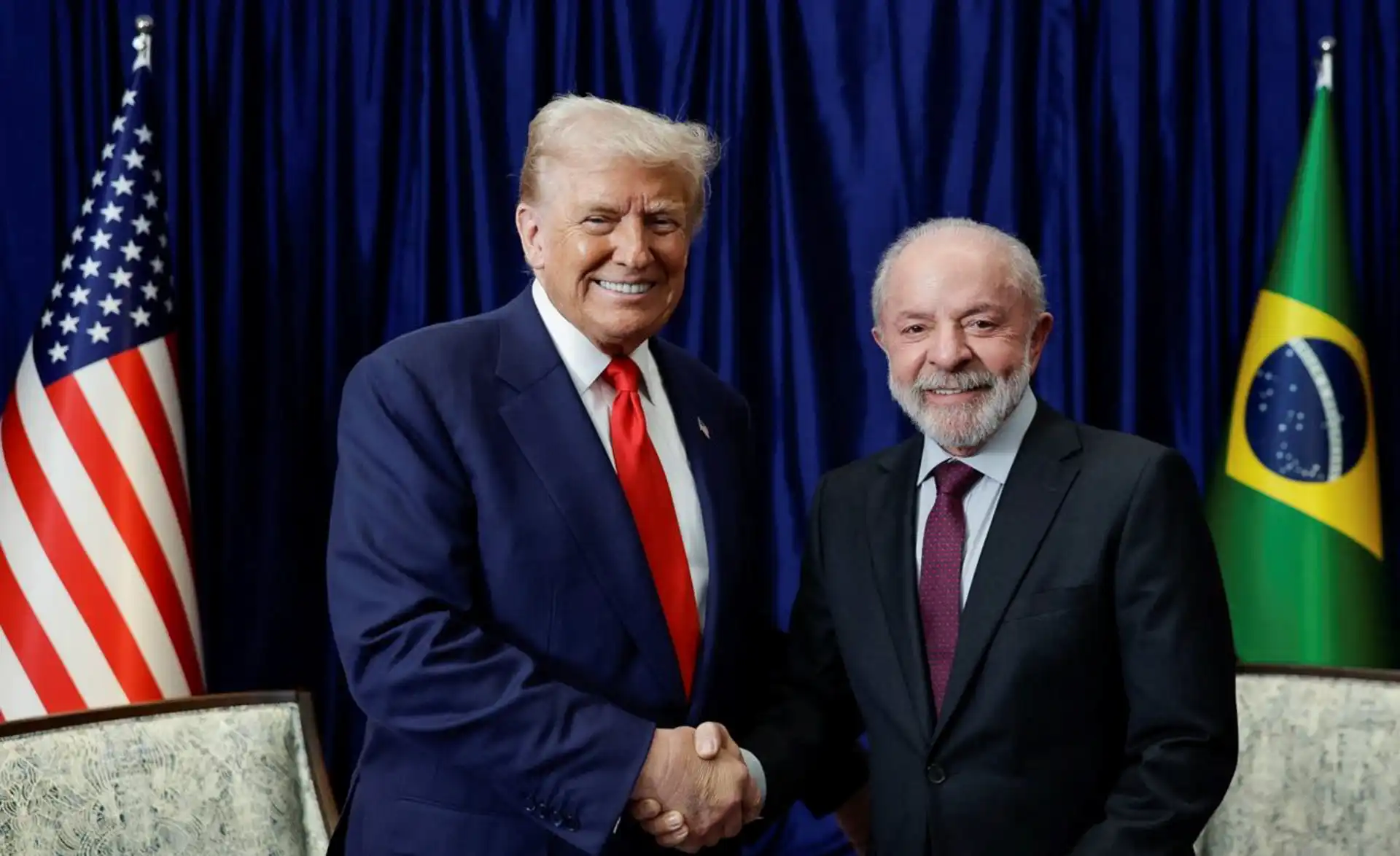 Imagem de Donald Trump e Luiz Inácio Lula da Silva sorrindo e apertando as mãos durante encontro oficial, com bandeiras dos Estados Unidos e Brasil ao fundo, simbolizando uma reunião entre líderes mundiais