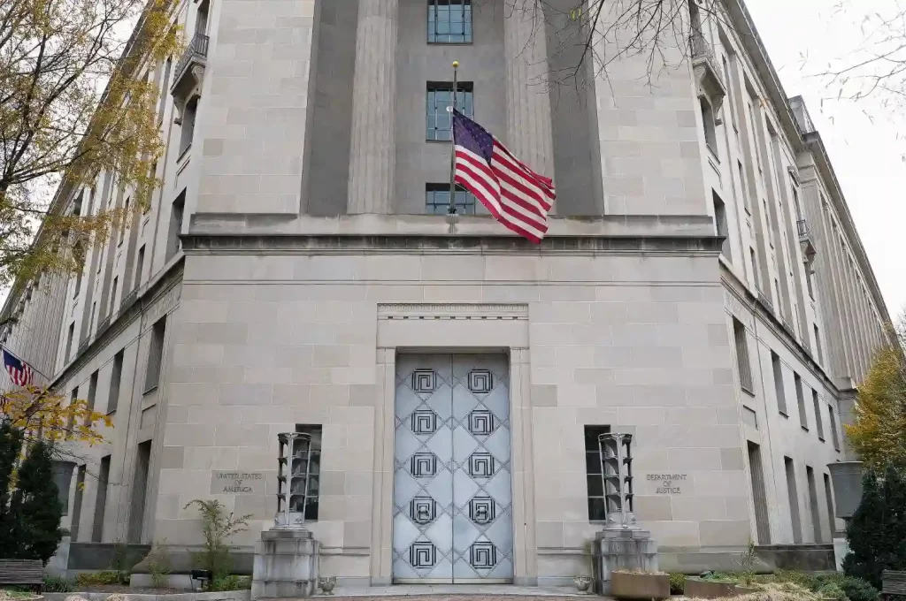 Imagem de um edifício público com destaque para a porta de entrada de metal, janelas e uma bandeira americana hasteada na fachada, simbolizando um órgão governamental ou edifício oficial nos Estados Unidos.