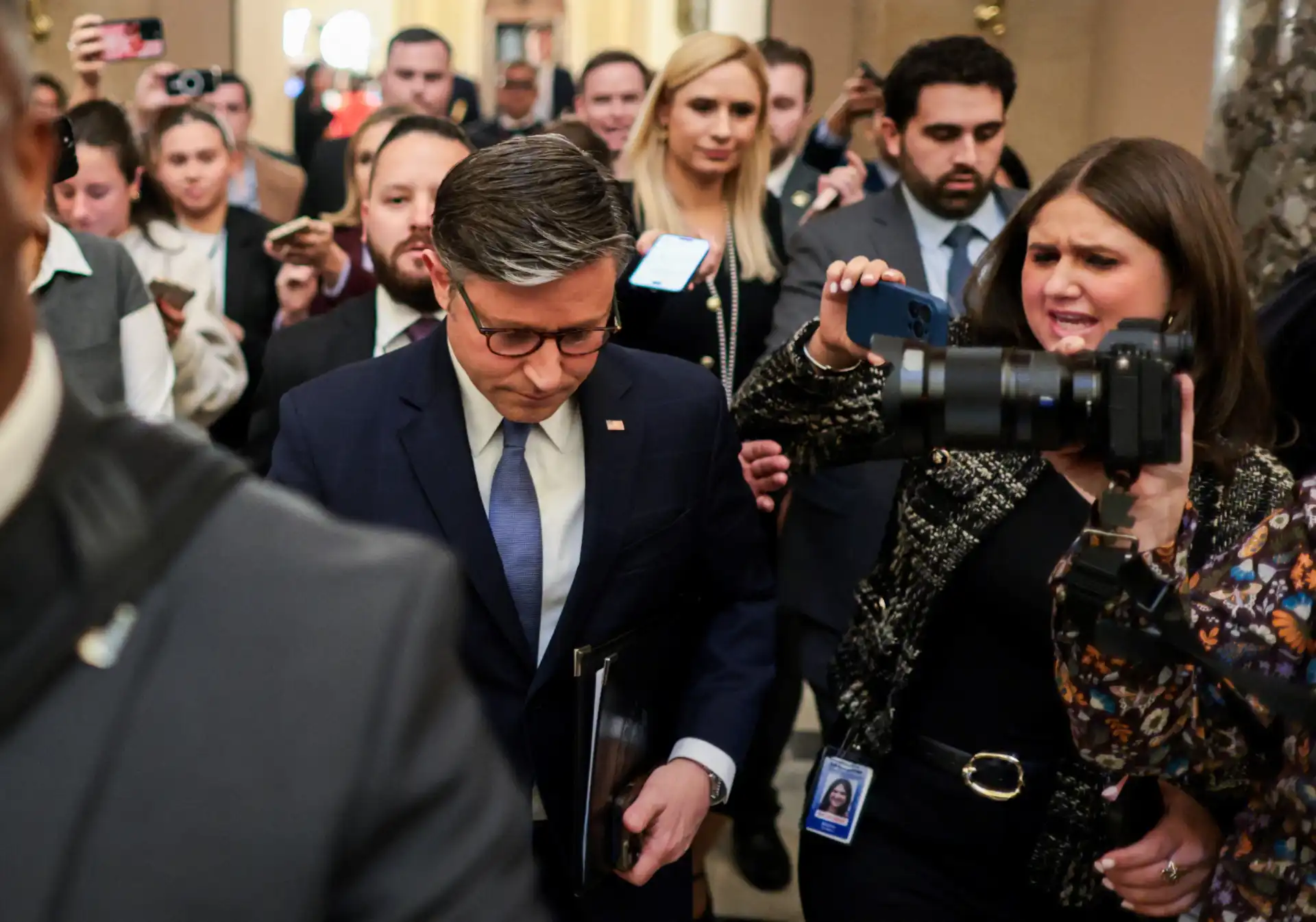 Imagem de um evento de imprensa com várias pessoas, incluindo um homem em terno escuro, ao centro, e uma mulher com câmera na mão, capturando o momento. O ambiente parece ser formal e agitado.