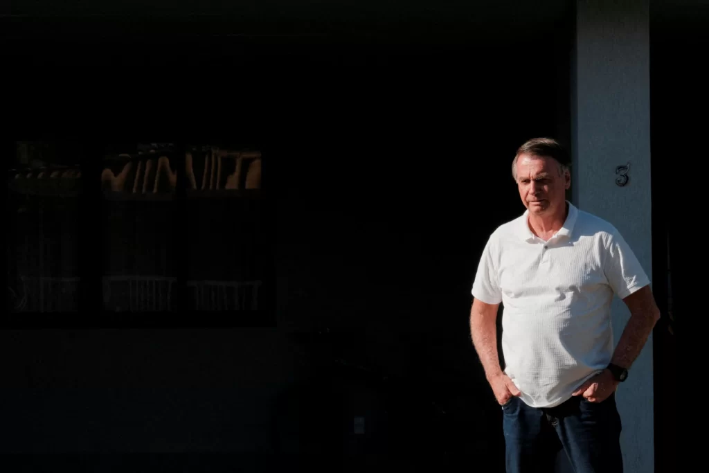 Homem de camiseta branca e calça jeans, com expressão séria, posando ao ar livre contra uma parede clara, em uma situação de iluminação com sombra.