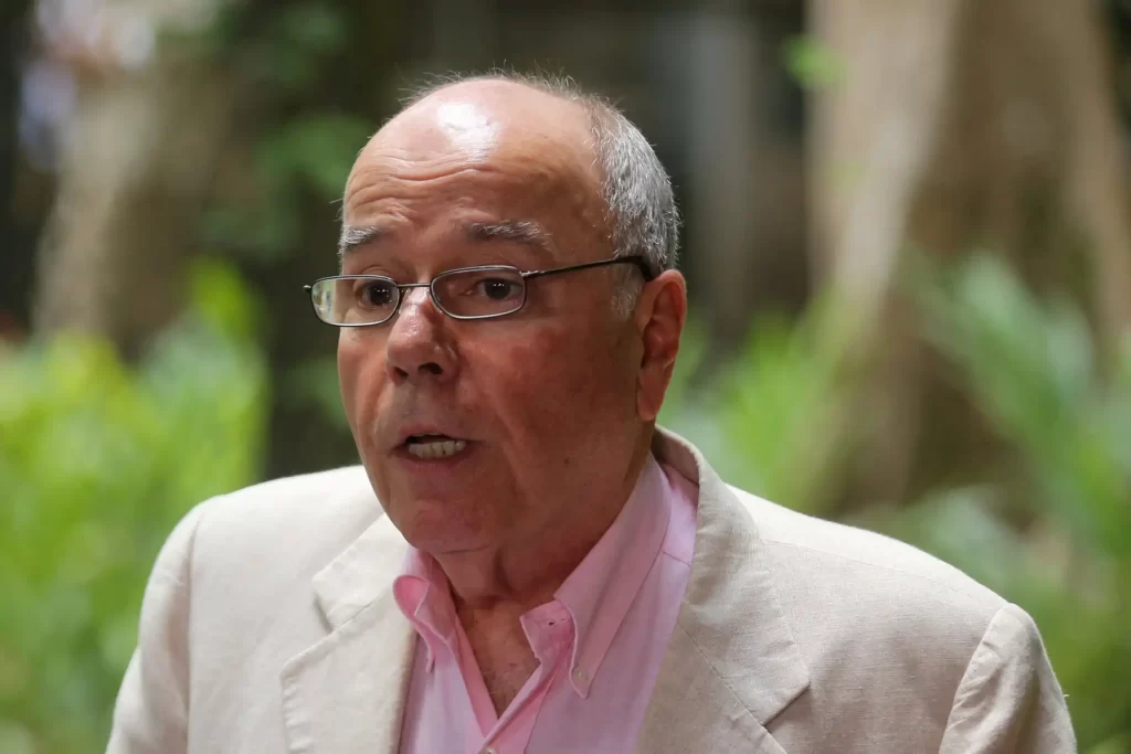 Homem idoso falando em ambiente natural ao ar livre, usando óculos e vestindo terno claro, na campanha ou evento ao ar livre.