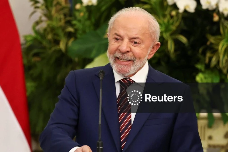 Homem idoso falando em evento formal, com terno e gravata, ao lado de uma bandeira, com fundo de plantas verdes.