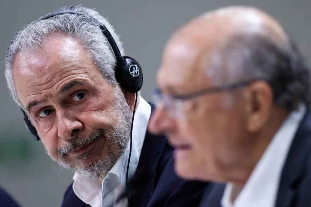 Homem com cabelo grisalho, barba, usando fone de ouvido, conversando com idoso de cabelos ralos, ambos em traje formal, em ambiente de reunião