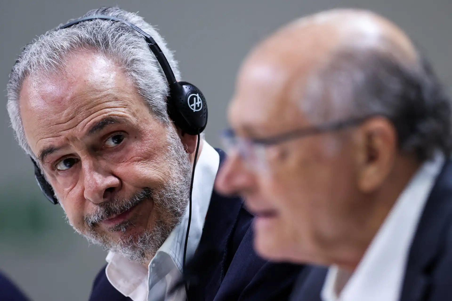 Homem com cabelo grisalho, barba, usando fone de ouvido, conversando com idoso de cabelos ralos, ambos em traje formal, em ambiente de reunião