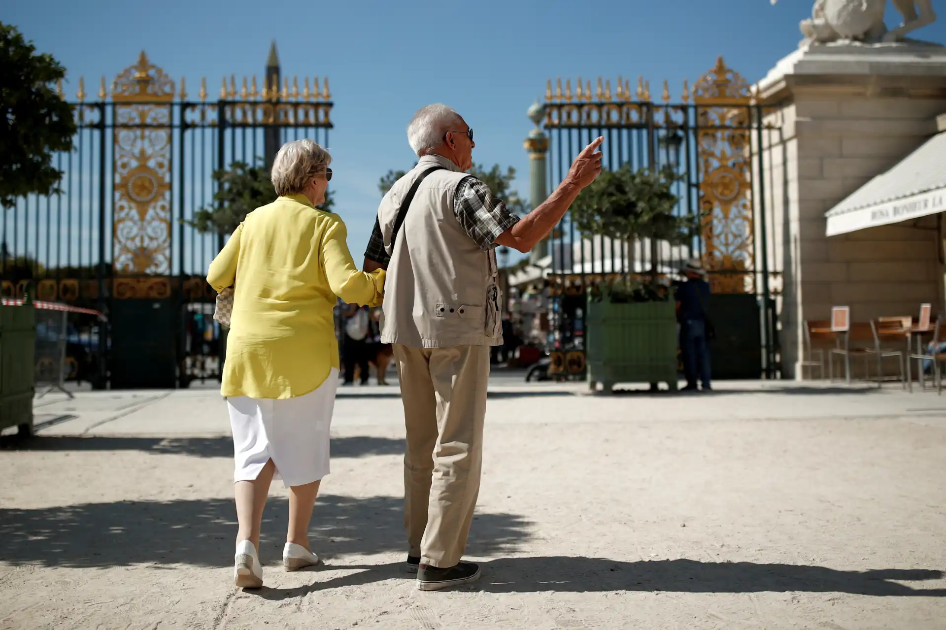 Idosos em passeio no parque com portão dourado ao fundo, aproveitando dia ensolarado.