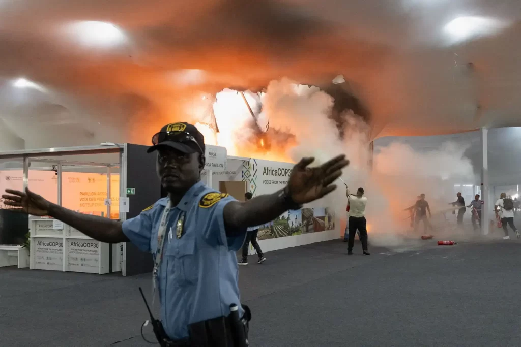 Pessoa de uniforme policial tentando controlar o caos durante um incêndio em uma feira ou evento na África, com fumaça e fogo ao fundo, em um ambiente de conflito.