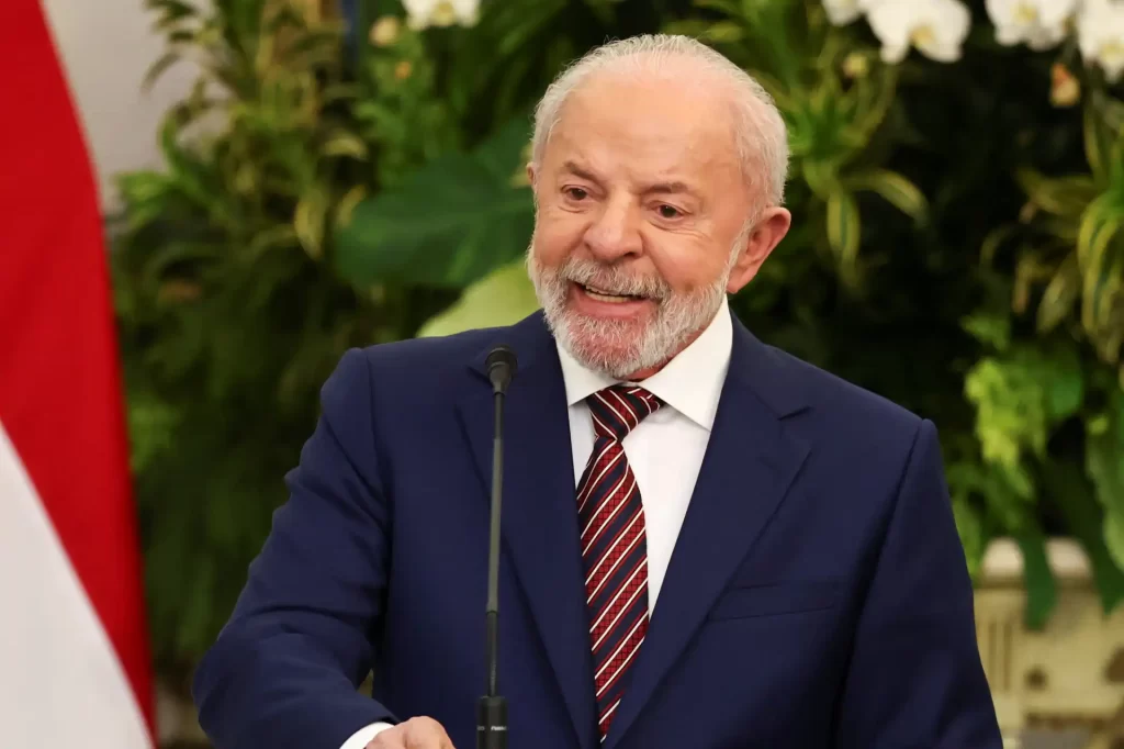 Homem sorridente com barba branca e cabelo grisalho, vestindo terno azul e gravata listrada, falando em evento com fundo de plantas verdes e bandeira do Brasil