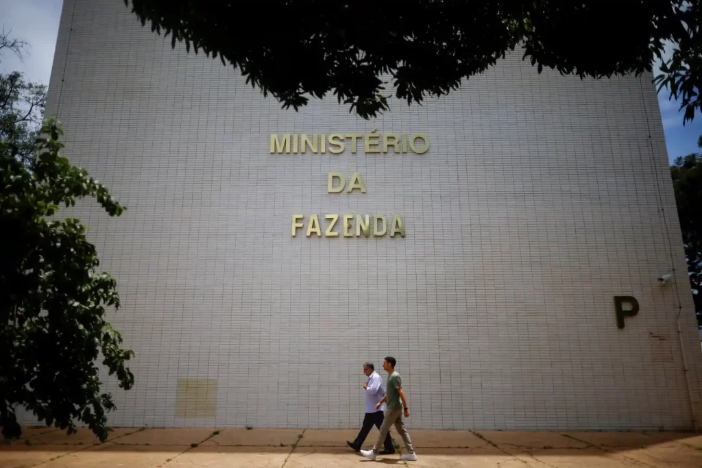 Edifício do Ministério da Fazenda com pessoas caminhando na calçada em frente, rodeado por árvores, em um dia claro.