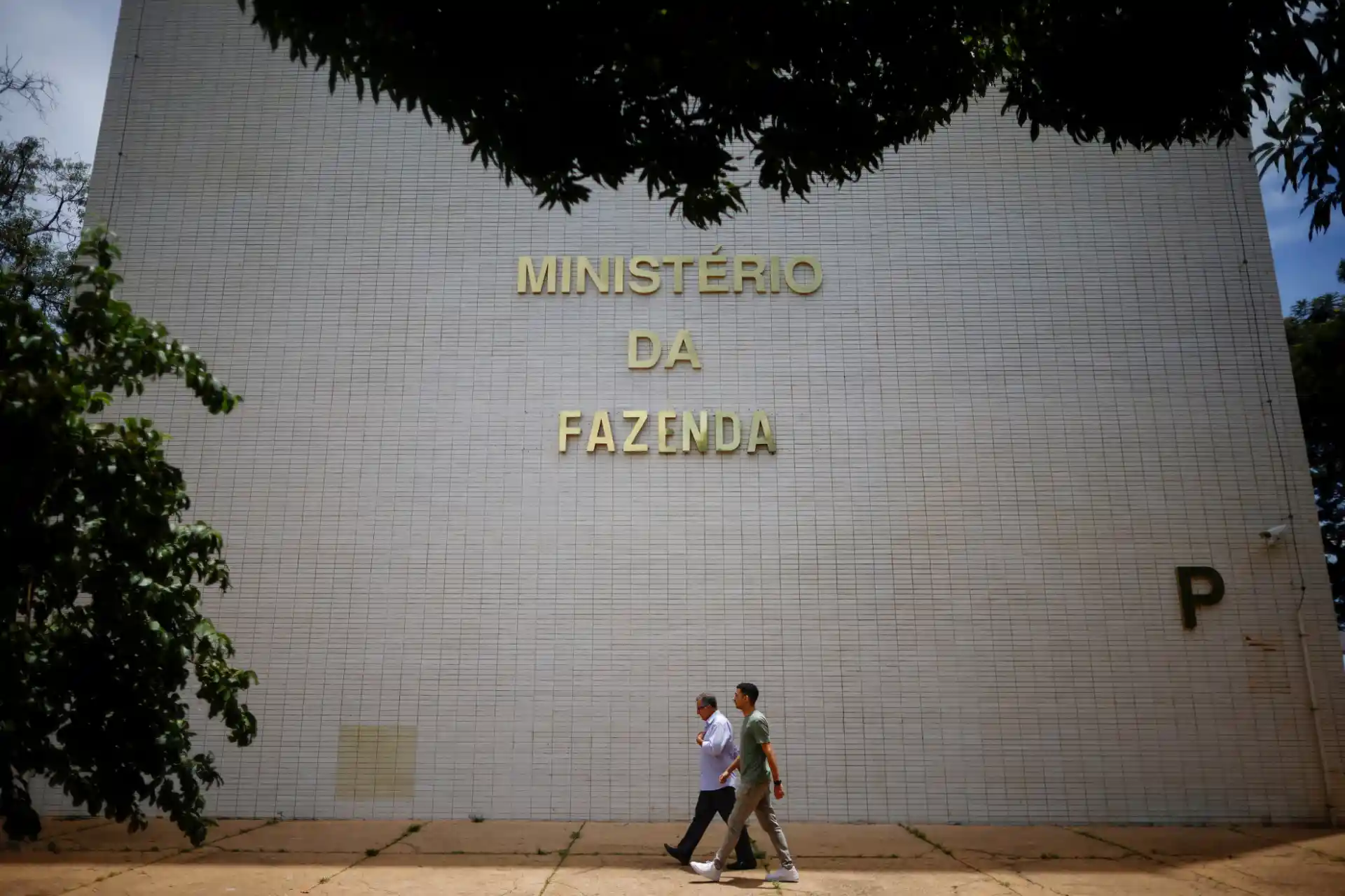 Edifício do Ministério da Fazenda com pessoas caminhando na calçada em frente, rodeado por árvores, em um dia claro.