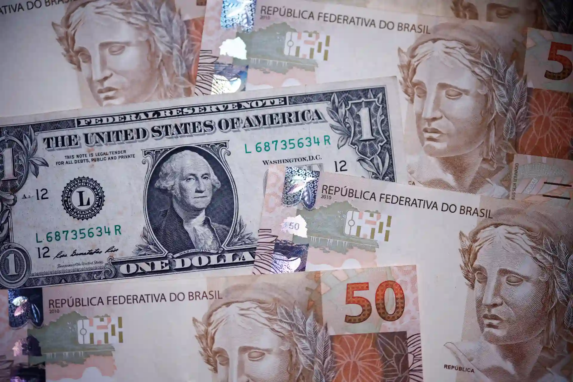 Imagem de várias cédulas de dinheiro, incluindo notas de 1 dólar americano e de 50 reais brasileiros, destacando as diferenças entre as moedas.