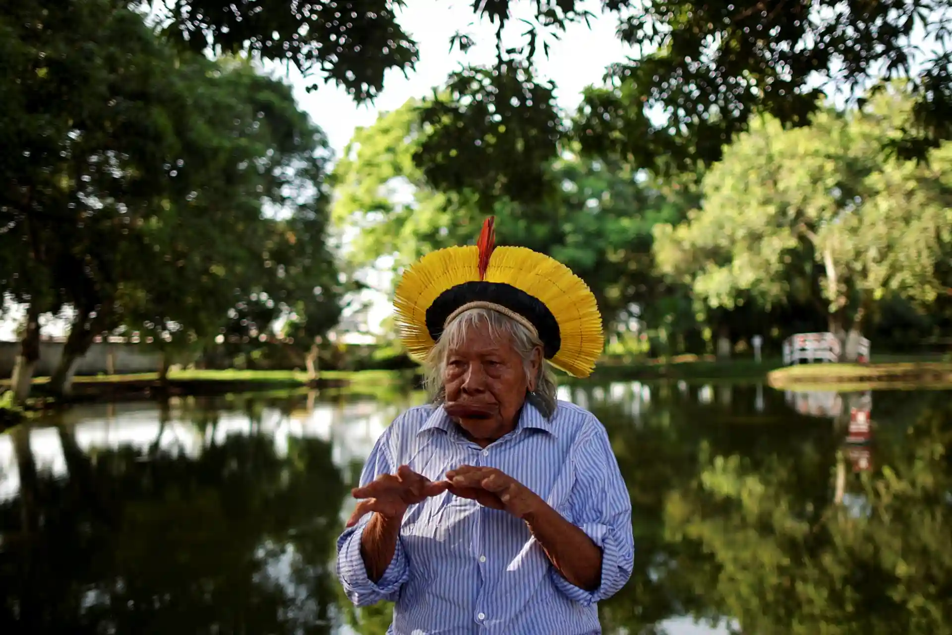 Mulher indígena com cabelo grisalho, usando um cocar colorido, ao ar livre na frente de um lago cercado de árvores verdes, em um ambiente natural e tranquilo.