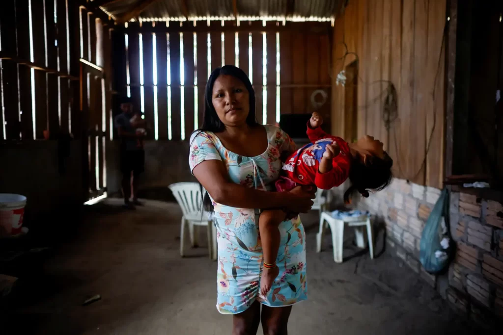 Mulher segurando uma criança dentro de uma casa de madeira, ambiente simples com móveis de plástico e iluminação natural,