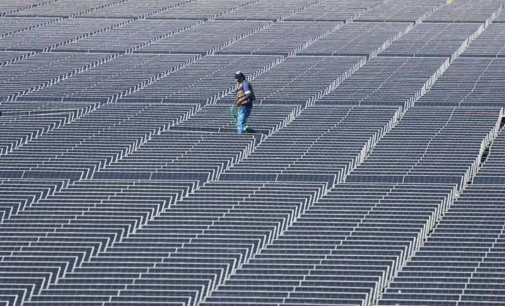 Pessoa caminhando por um vasto campo de painéis solares dispostos em linhas ordenadas ao ar livre, aproveitando a energia solar.