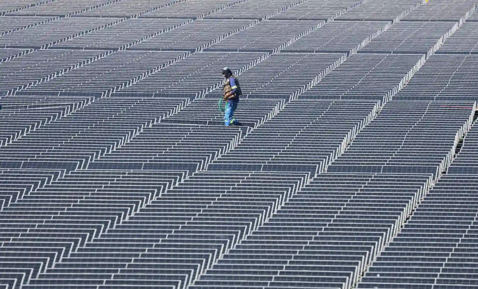Pessoa caminhando por um vasto campo de painéis solares dispostos em linhas ordenadas ao ar livre, aproveitando a energia solar.