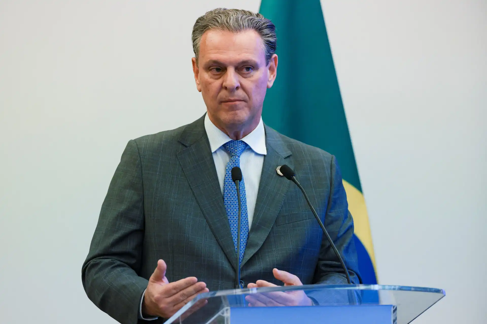 Político brasileiro em discurso na frente de uma bandeira, vestindo terno e gravata, em um evento oficial.