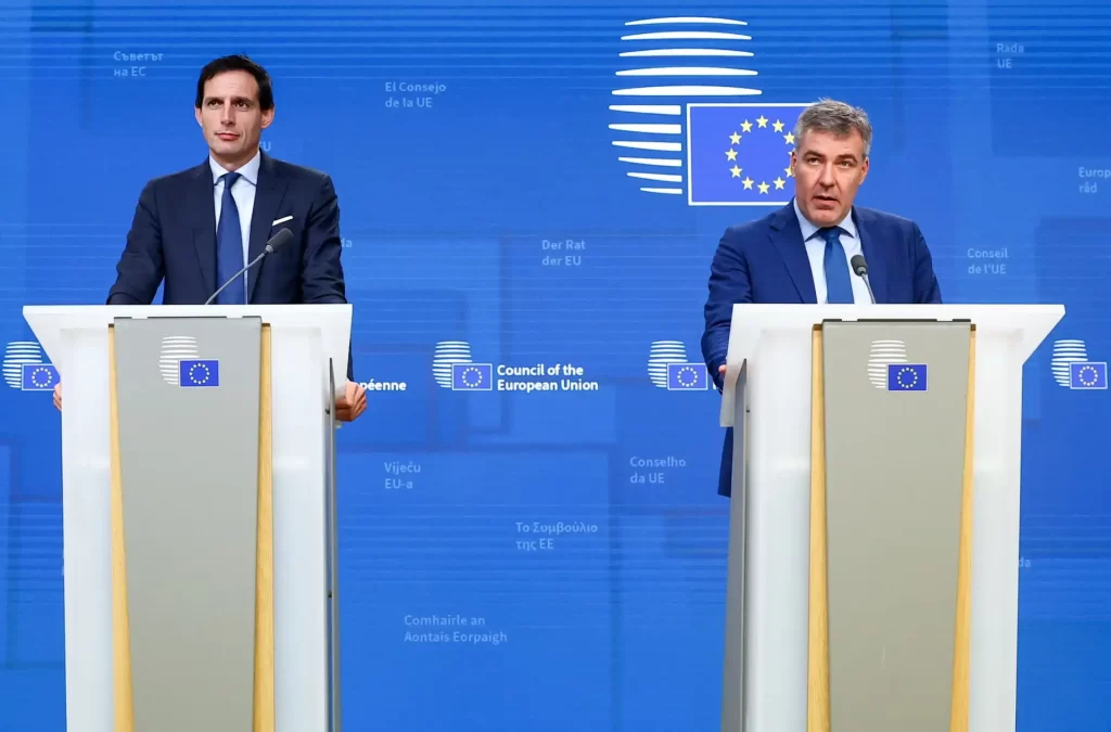 Dois homens em pé, falando em uma coletiva de imprensa na União Europeia, com um fundo azul com o logo da UE e referências às instituições europeias.