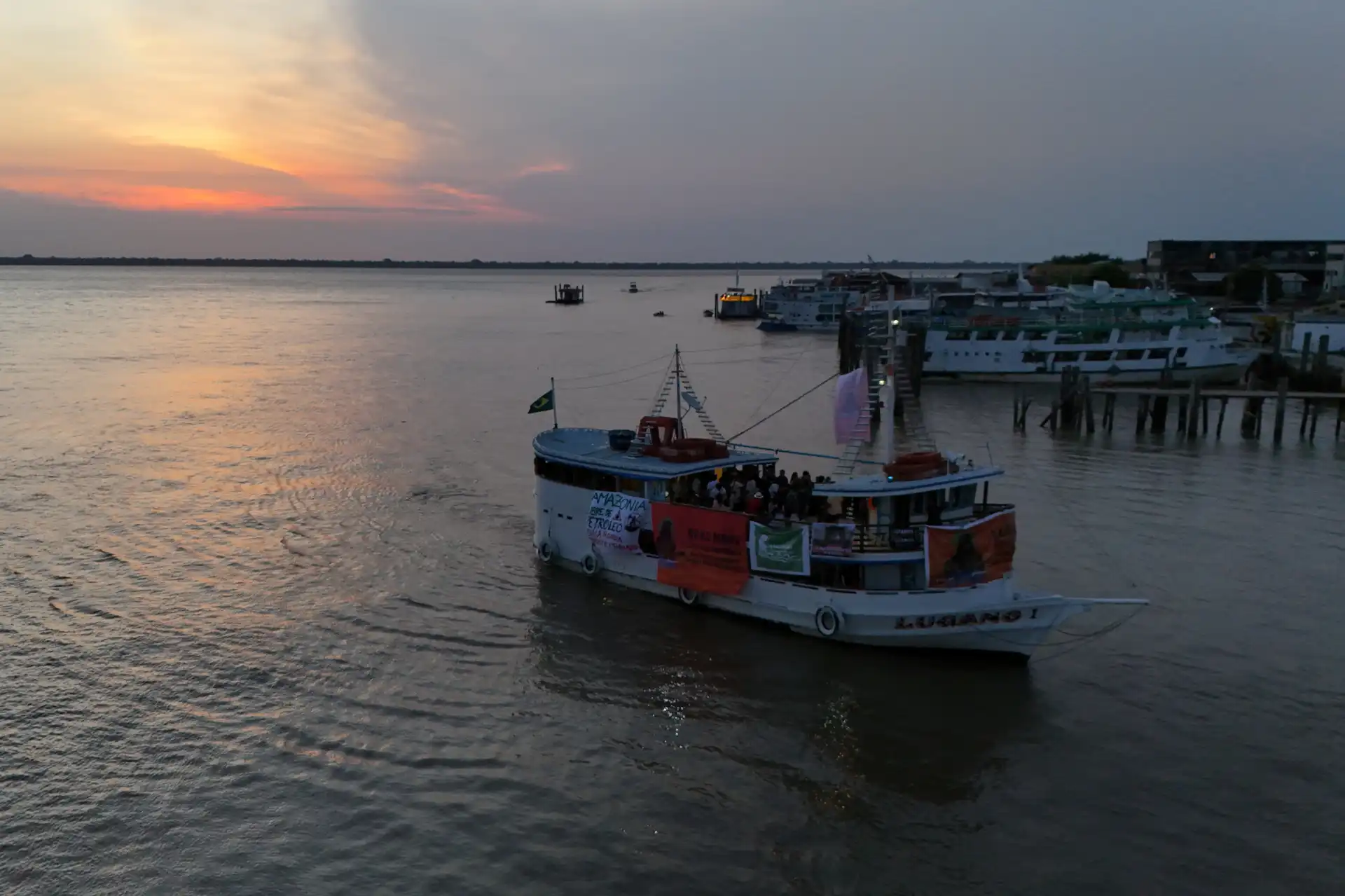 Pôr do sol no rio com barcos atracados e uma embarcação se deslocando, cenário tranquilo e agradável no final da tarde com céu colorido e água calma.