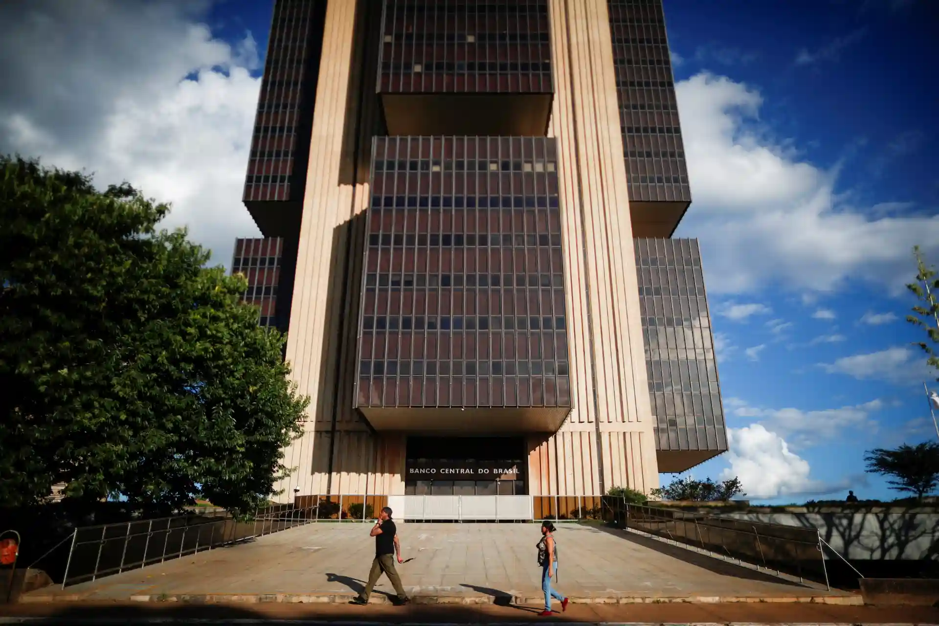 Imagem do edifício do Banco Central do Brasil, uma construção moderna de grande altura com fachada de vidro e concreto, localizado em Brasília, sob céu claro com nuvens.