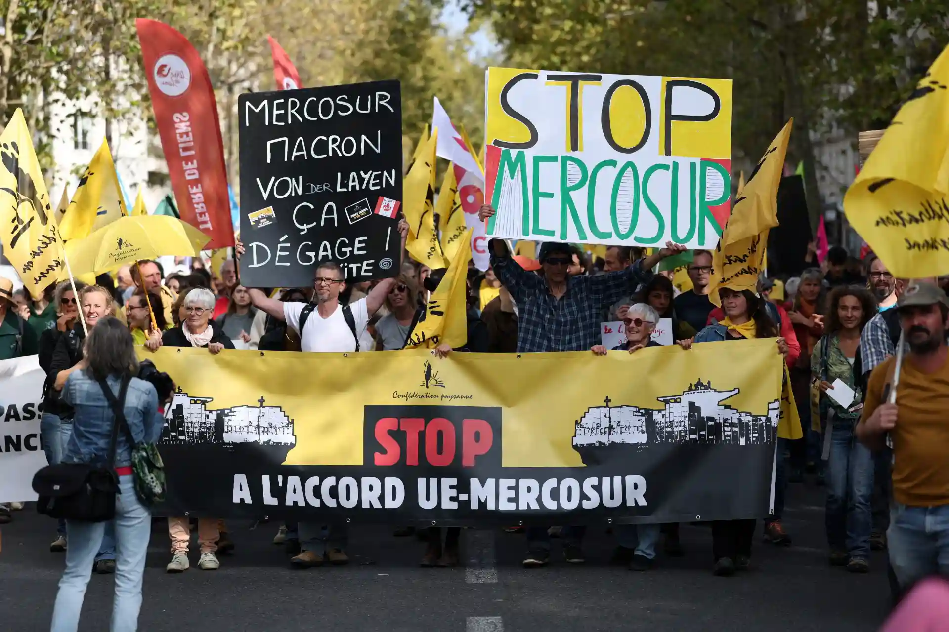 Manifestantes protestam contra o acordo UE-MERCOSUR em uma manifestação na rua, segurando faixas e cartazes, incluindo mensagens como 'Stop Mercosur' e 'Mercosur Macron'.