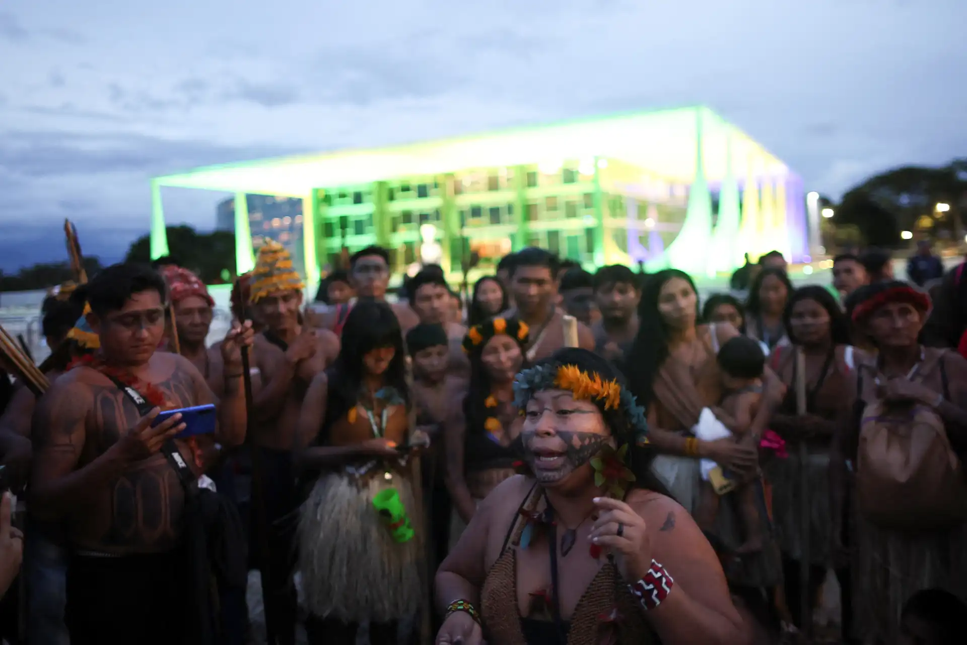 Grupo de pessoas participando de uma celebração cultural ao ar livre, com uma estrutura iluminada ao fundo, durante o entardecer, em uma festa tradicional.