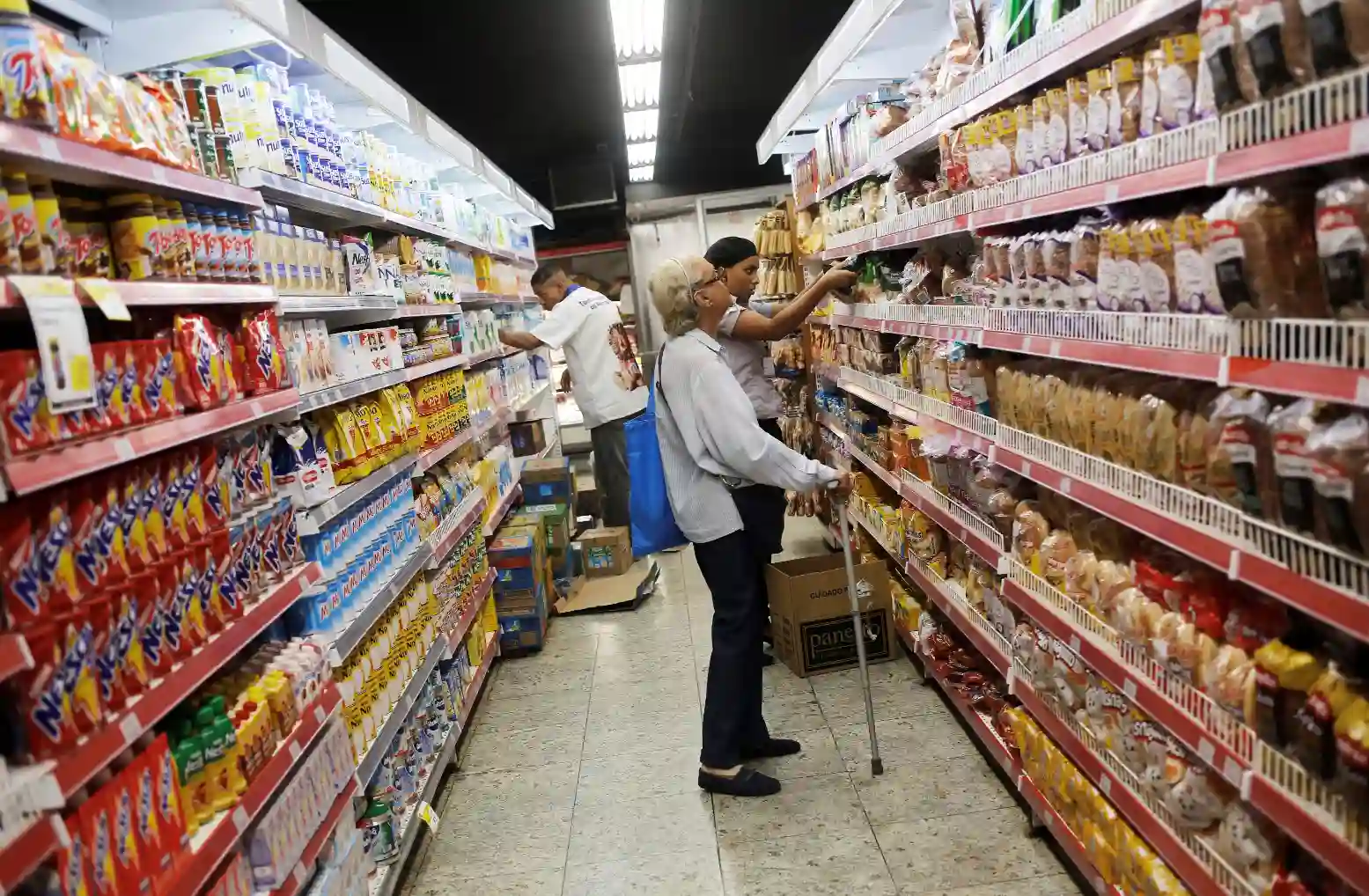 Crianças e idosos fazendo compras em supermercado com várias opções de produtos alimentícios nas prateleiras.