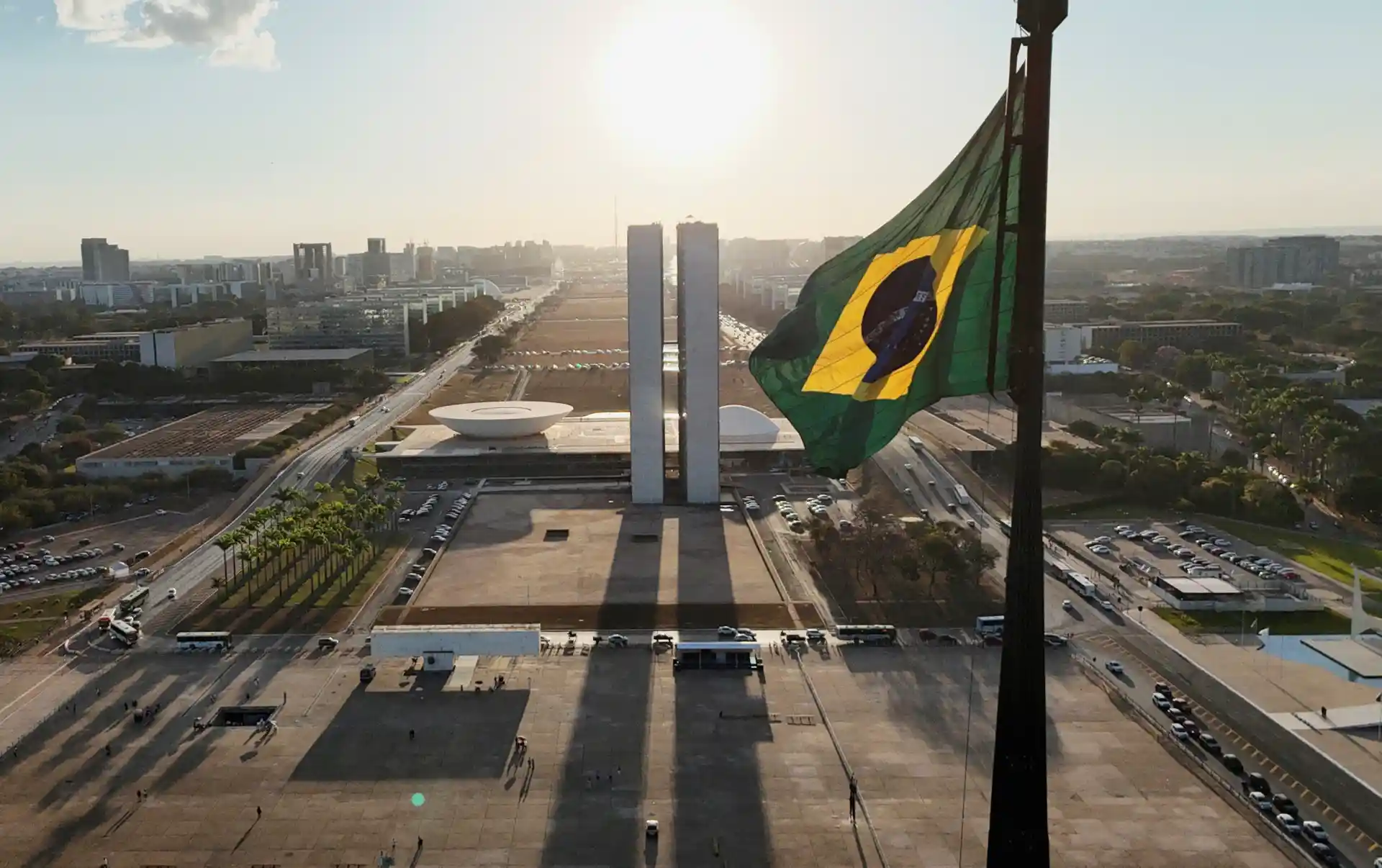 Vista aérea do Congresso Nacional em Brasília com a bandeira do Brasil ao vento, capturada ao pôr do sol, destacando a importância do símbolo nacional.