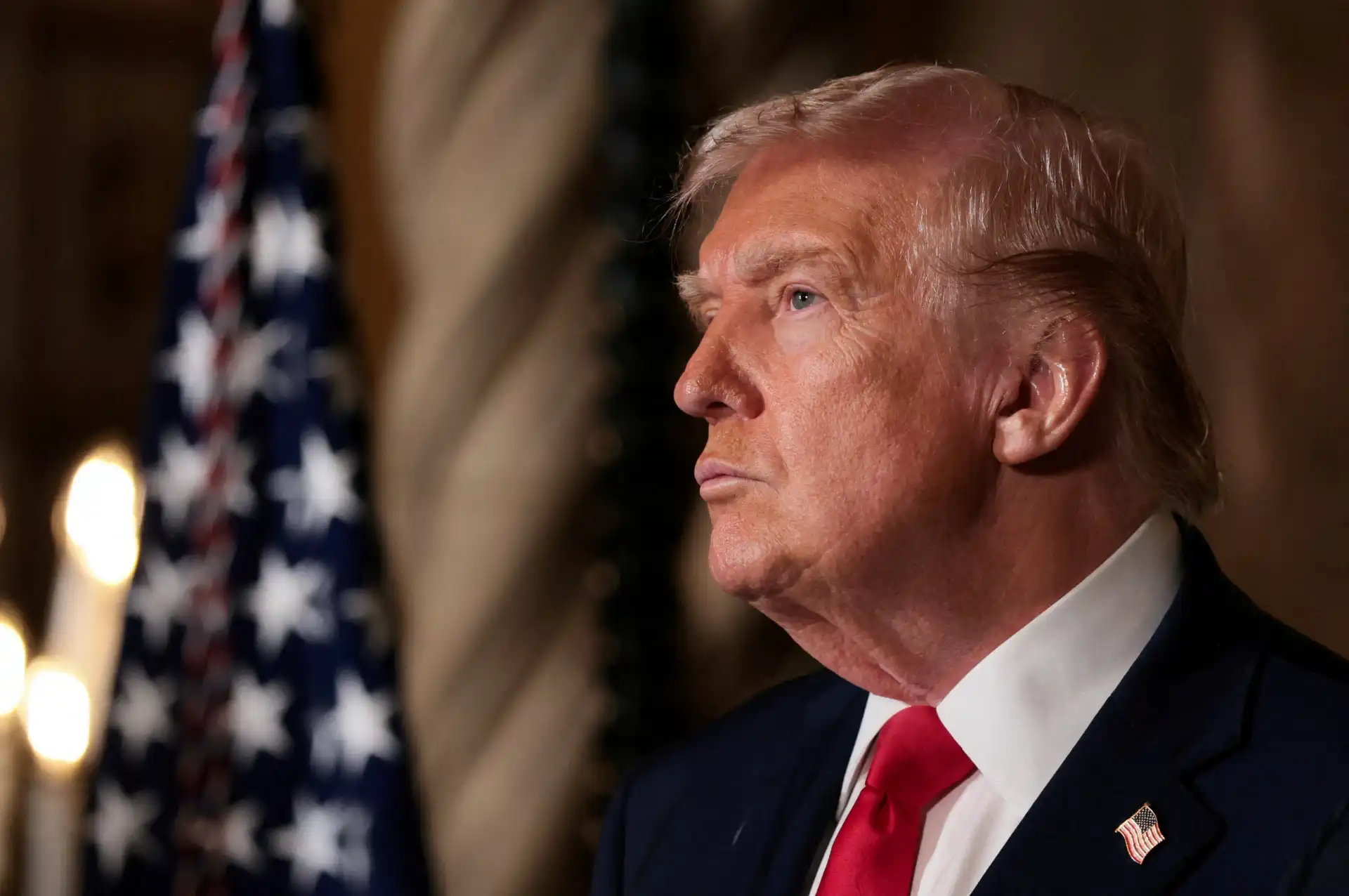 Imagem de Donald Trump em perfil, com fundo de bandeira americana e decoração clássica, durante evento oficial. Foco na expressão séria e postura de liderança do ex-presidente dos Estados Unidos.