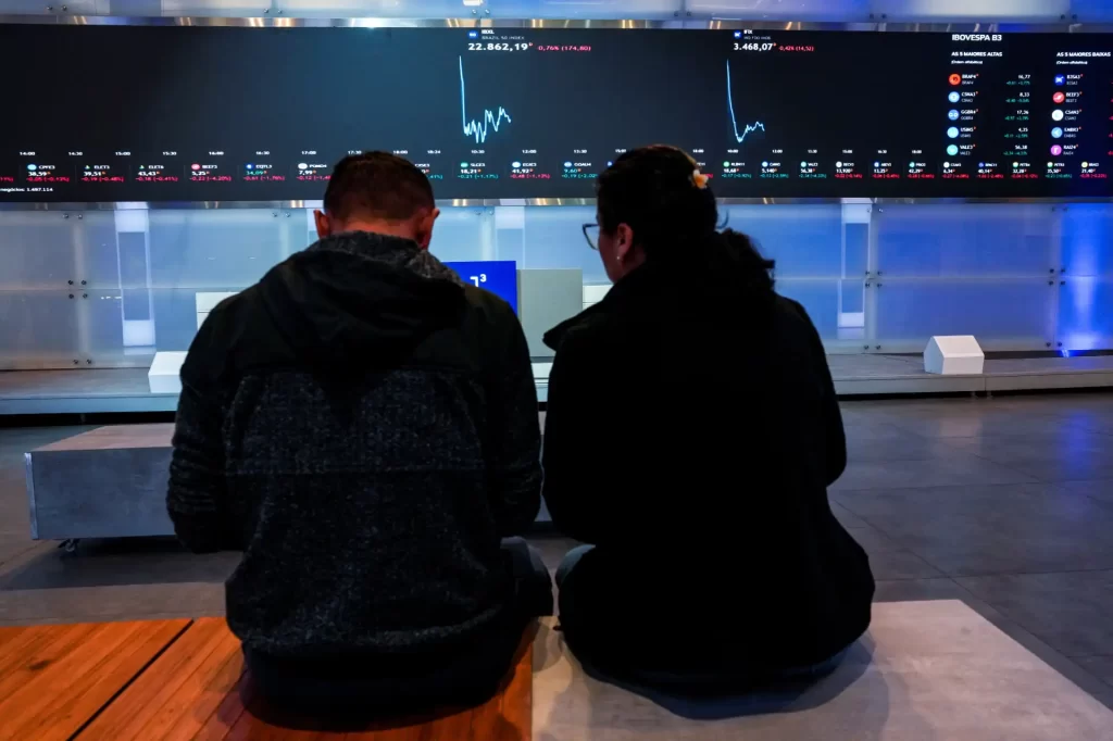 Dupla de pessoas observando gráficos e dados financeiros em uma tela de monitoramento na bolsa de valores, destacando análises de mercado financeiro.