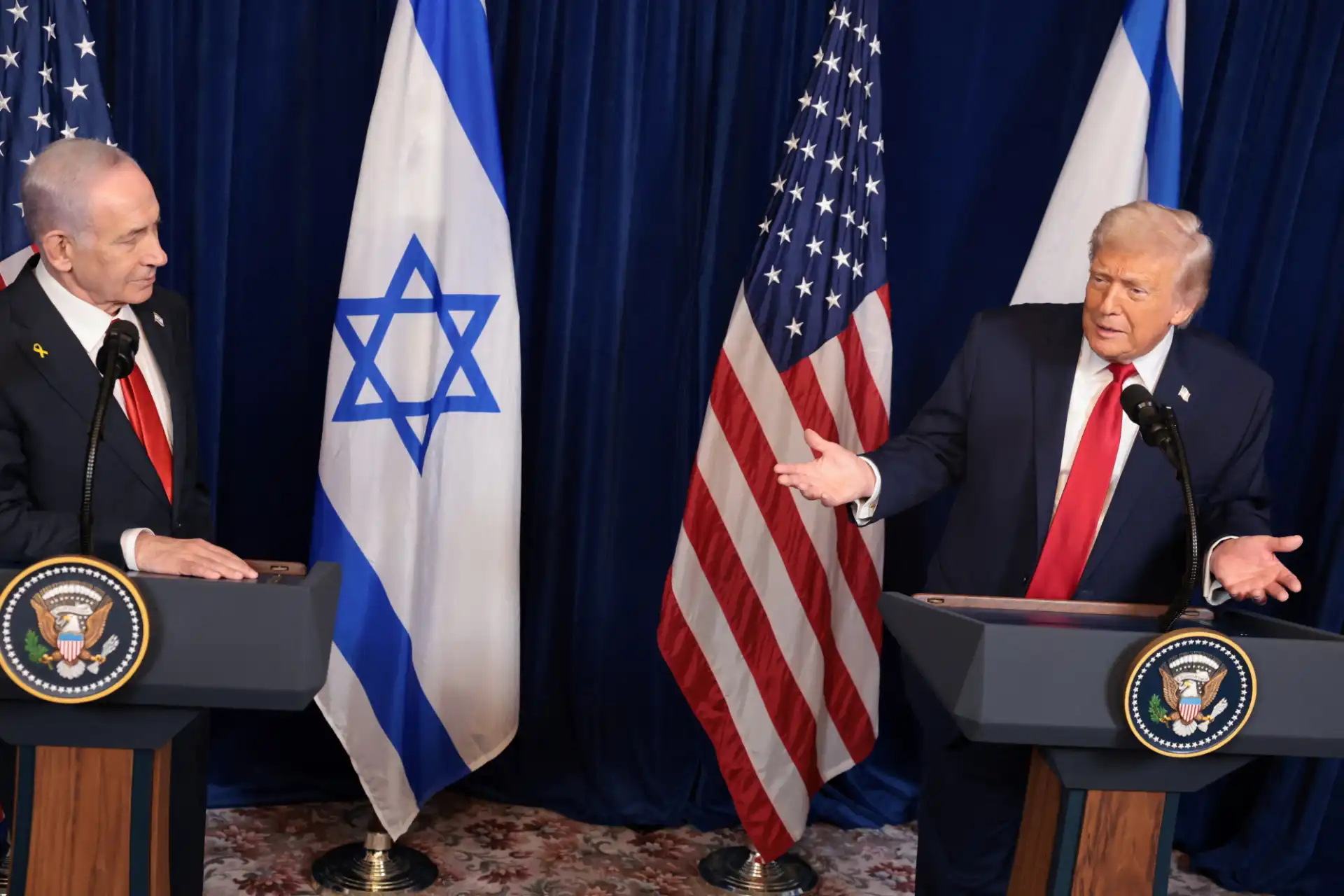 Imagem de um encontro entre o Primeiro Ministro de Israel e o ex-presidente dos EUA, Donald Trump, em uma entrevista ou conferência, com bandeiras de Israel e Estados Unidos ao fundo.