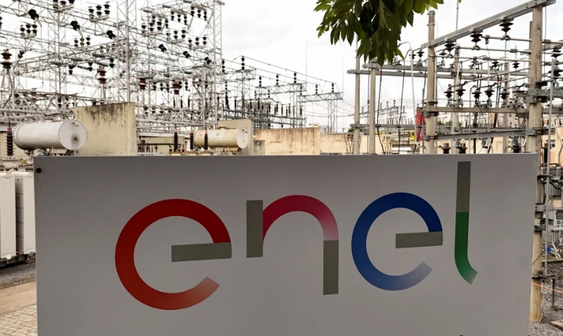 Placa com a palavra 'Enel' em destaque, indicando uma fonte de energia elétrica, instalada em frente a uma subestação de energia com estruturas de alta tensão ao fundo.