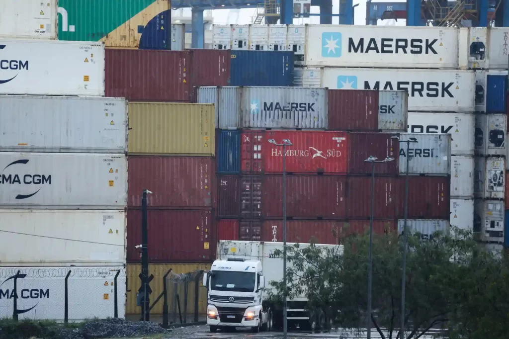 Estoque de containers de transporte marítimo em um porto, destacando marcas como Maersk e Hamburg Süd, com caminhões ao fundo.