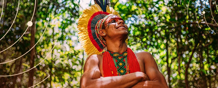 Homem indígena usando roupas tradicionais e ornamentos em um ambiente natural com árvores e folhagem, sorrindo com orgulho e felicidade. Marco Temporal
