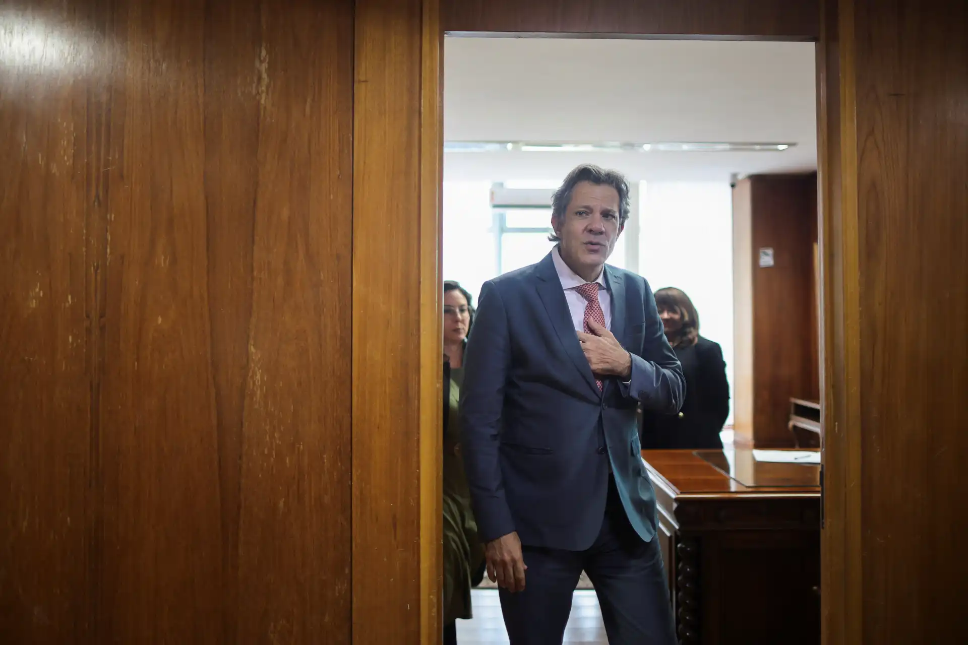 Homem de terno azul e gravata no interior de um edifício, segurando a mão no peito, visível entre duas paredes de madeira, em uma ocasião formal.