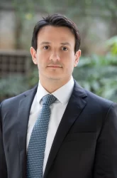 Homem de terno apresentando uma expressão séria e confiante, com cabelo escuro, em ambiente ao ar livre com fundo de árvores e folhagens.