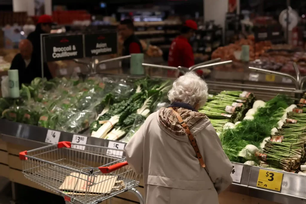 Idosa fazendo compras de hortifrúti em supermercado, observando vegetais frescos na seção de verduras e legumes.