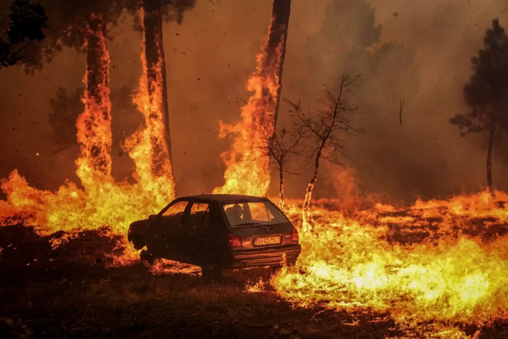Carro destruído em meio a um incêndio florestal devastador, com chamas altas e fumaça escura em uma floresta queimando intensamente.