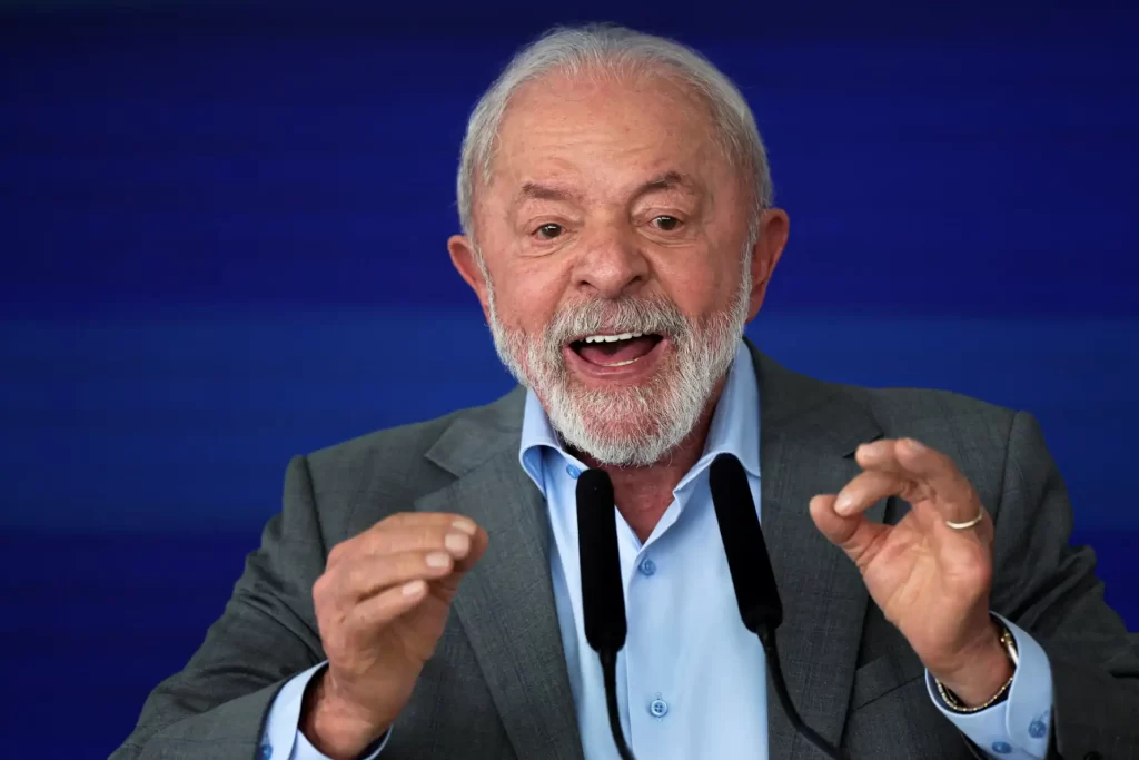 Lula da Silva, ex-presidente do Brasil, falando em público com entusiasmo, usando terno cinza e camisa azul, expressão empolgada, fundo azul.