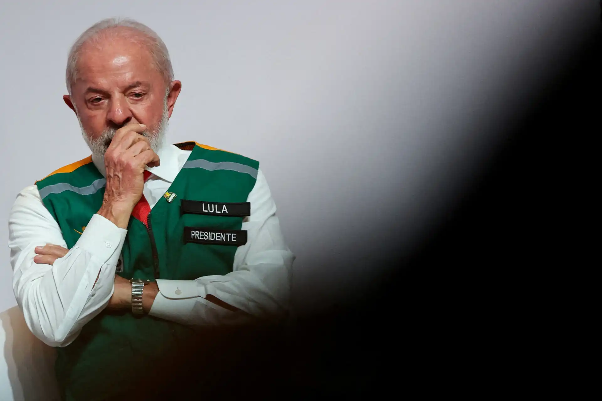 Imagem de Lula, ex-presidente do Brasil, trajando colete com os dizeres 'Lula' e 'Presidente', com expressão pensativa. Fundo com efeito de luz e sombra.