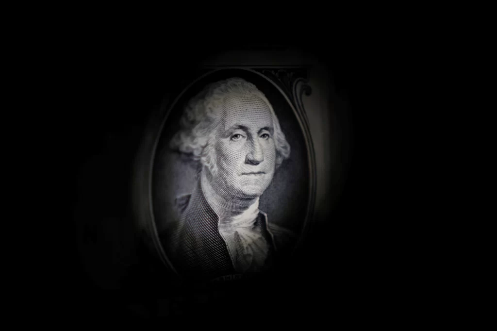 Imagem de uma nota de dólar com retrato de Benjamin Franklin, destacado em iluminação escura para foco no rosto e detalhes.