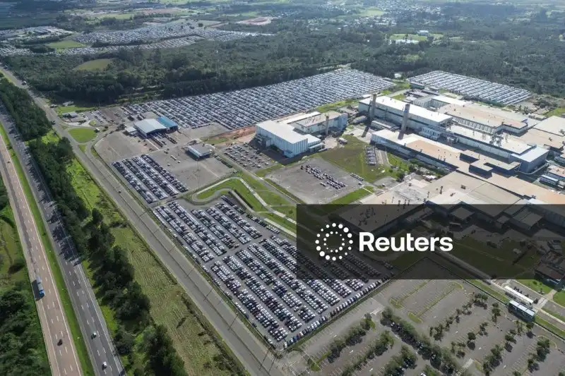 Imagem aérea de um grande parque de estacionamento ao redor de uma industrial, com áreas de acesso de veículos e uma estrada ao lado, em meio a uma área verde.