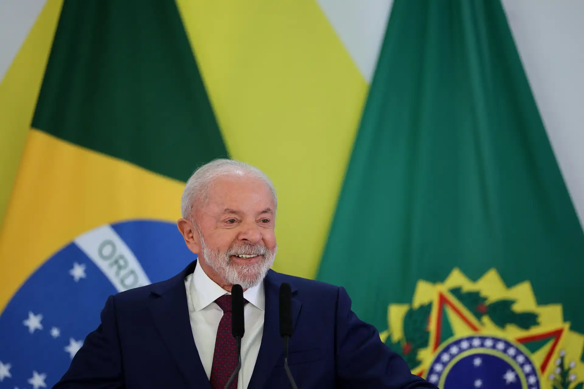 O presidente falando durante uma cerimônia, com bandeiras do Brasil ao fundo. A imagem destaca sua expressão confiante e sorridente, sendo uma cena oficial.