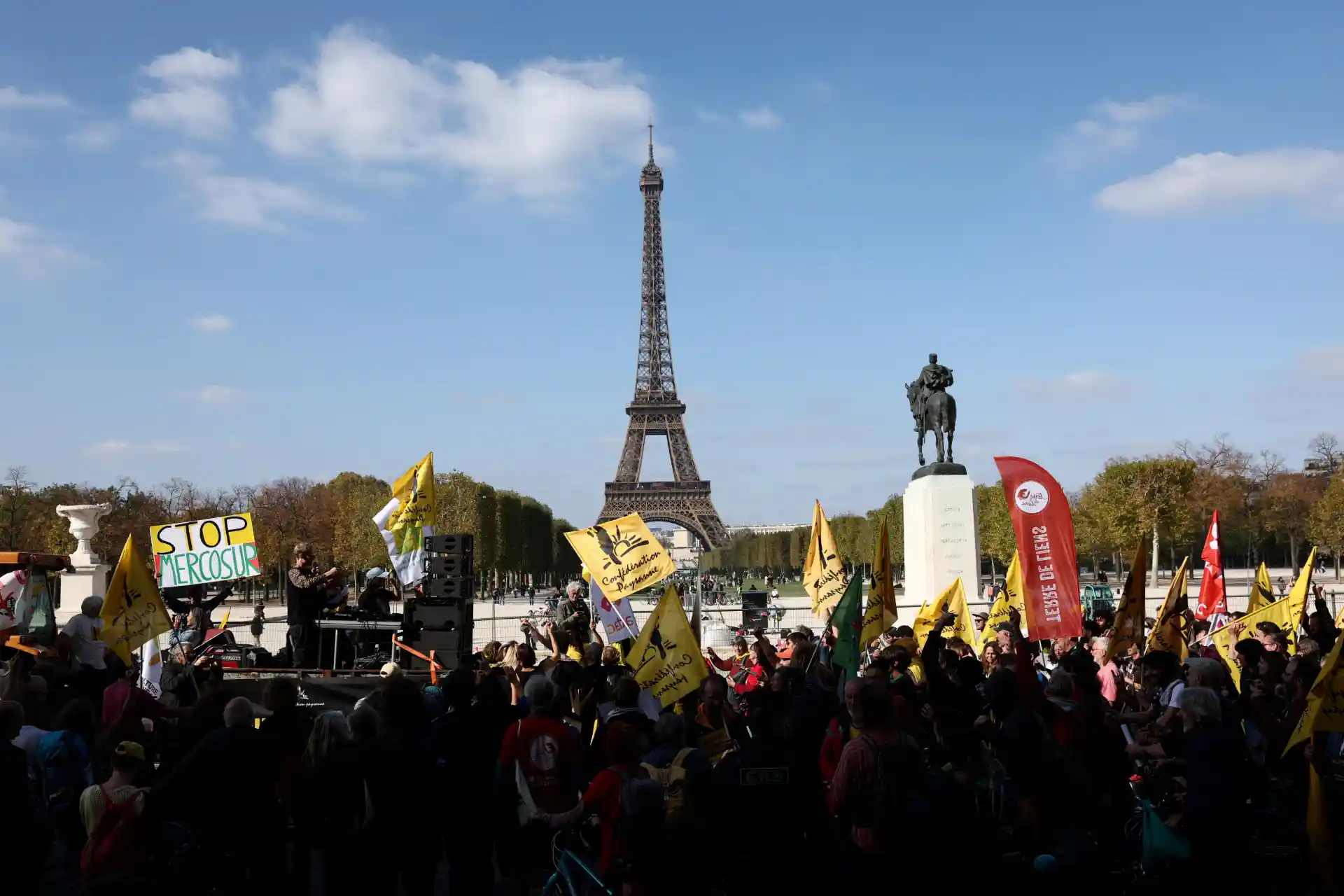 Protesto na Praça do Trocadero em frente à Torre Eiffel, em Paris, com manifestantes segurando bandeiras e cartazes, incluindo um com a mensagem "Stop Mergosur".