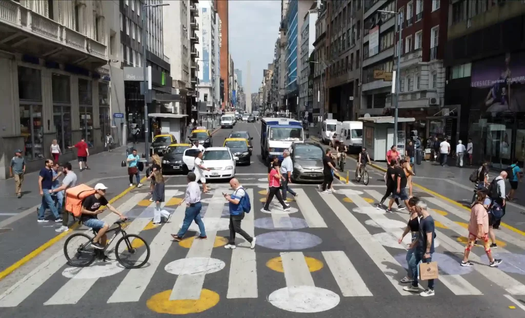 Rua movimentada com pedestres e ciclistas cruzando em uma cidade grande, rodeada por edifícios comerciais e escritórios, durante o dia.