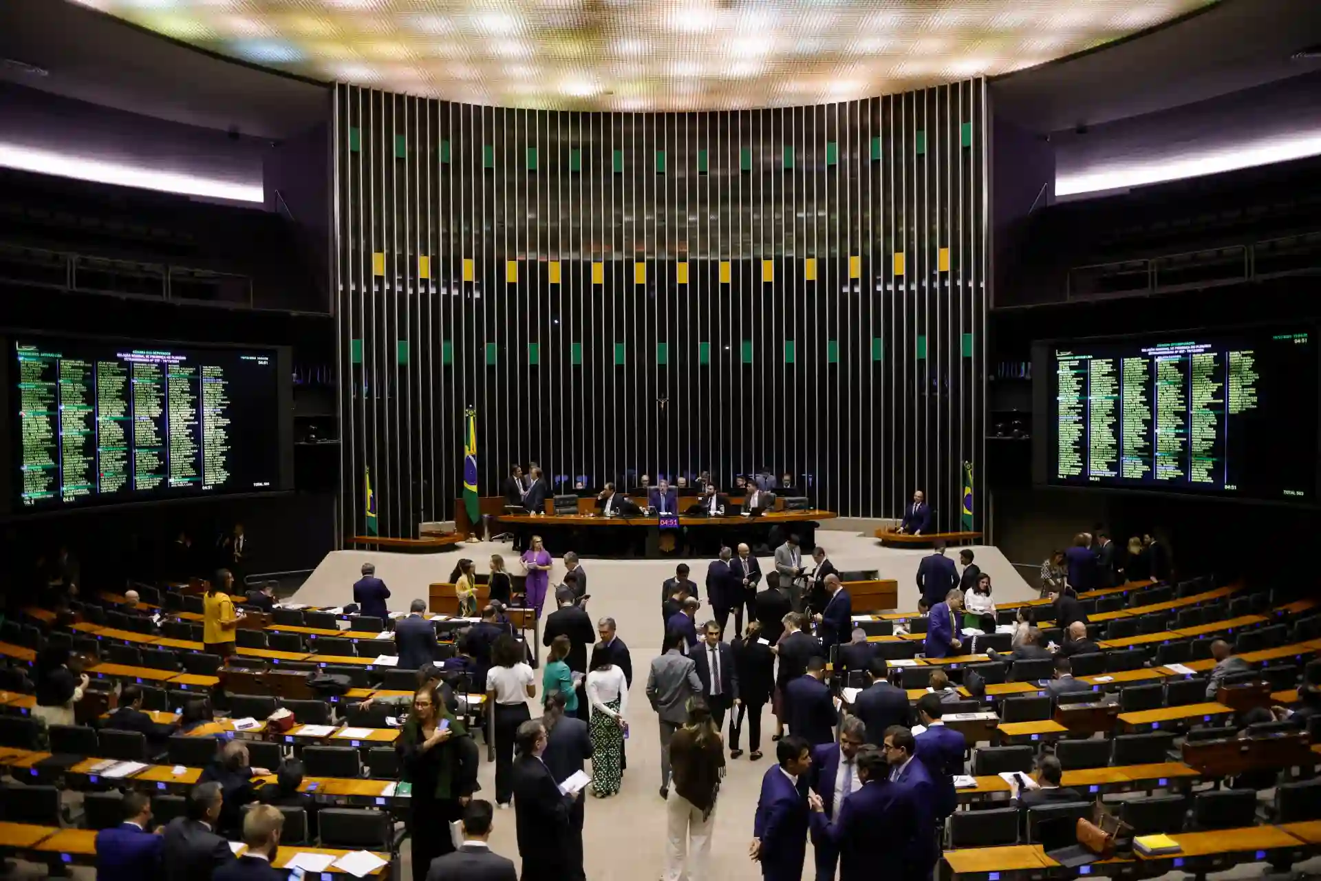 Sessão plenária no parlamento brasileiro com deputados e senadores discutindo em harmonia, com destaque para a bandeira do Brasil e telas informativas ao fundo.