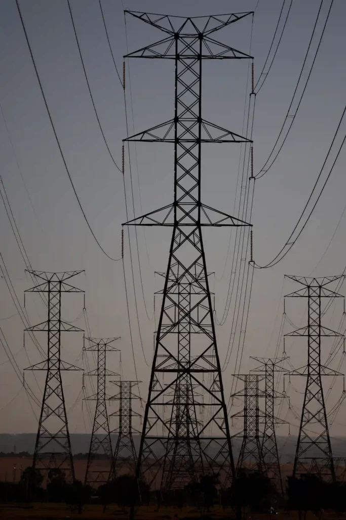 Imagem de várias torres de transmissão de energia elétrica com linhas de alta tensão contra o céu ao nascer ou por do sol. Destaque para uma torre maior ao centro e várias menores ao redor, mostrando a infraestrutura de distribuição de energia elétrica.