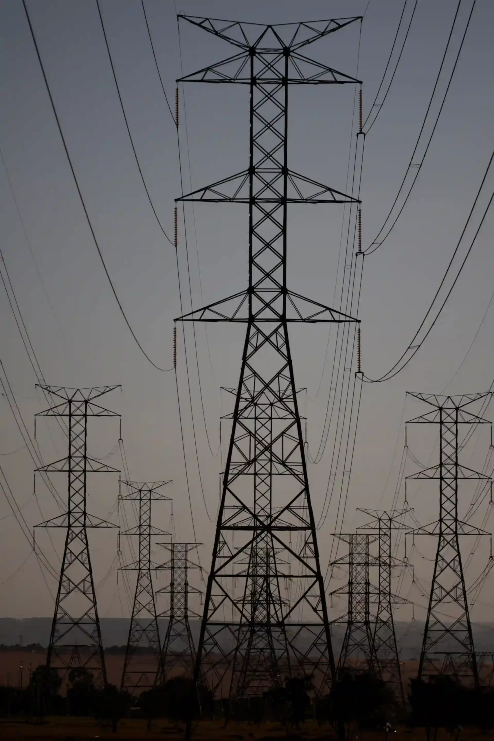 Imagem de várias torres de transmissão de energia elétrica com linhas de alta tensão contra o céu ao nascer ou por do sol. Destaque para uma torre maior ao centro e várias menores ao redor, mostrando a infraestrutura de distribuição de energia elétrica.