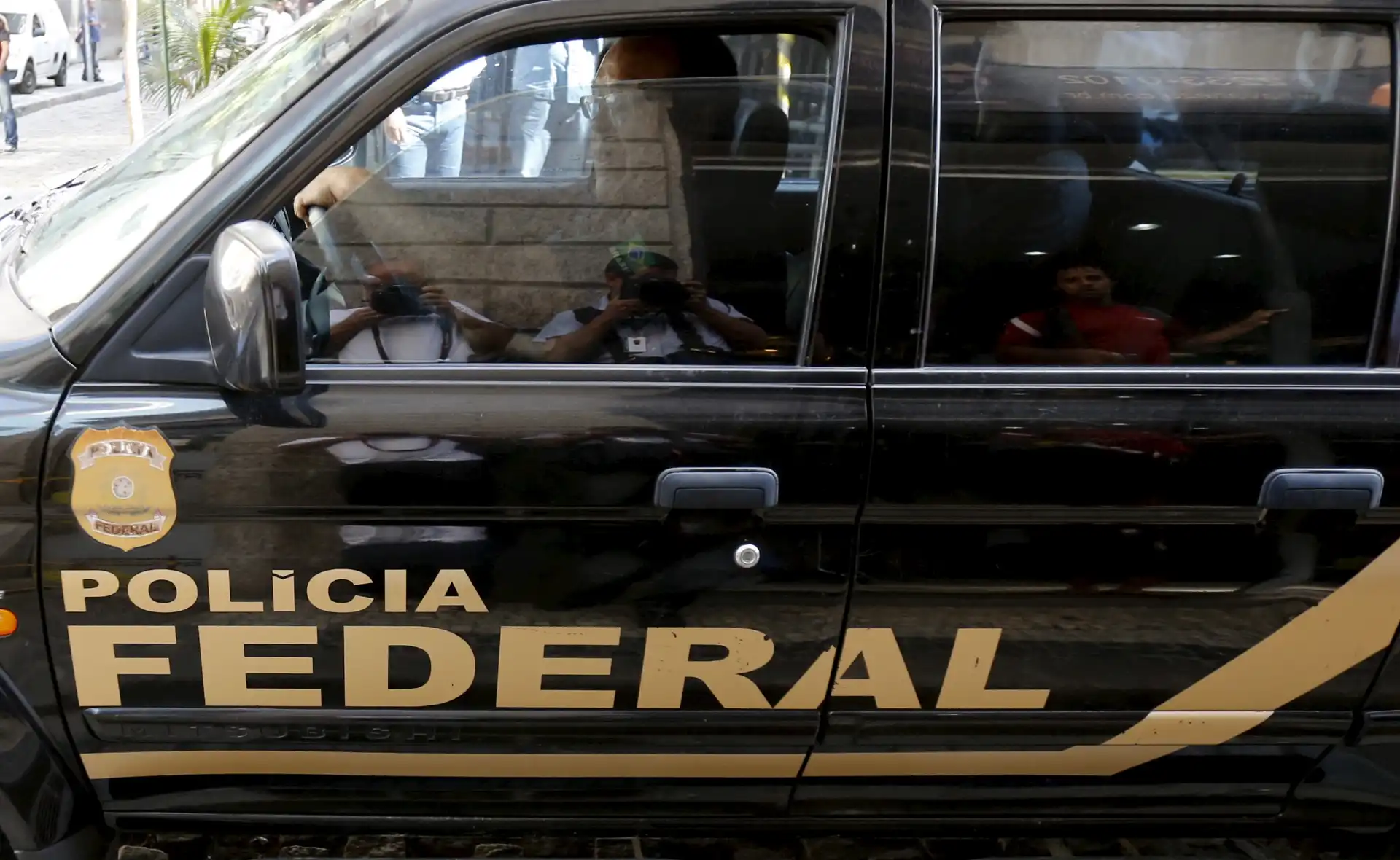 Veículo oficial da Polícia Federal preto com detalhes dourados, estacionamento em área urbana, refletindo pessoas no vidro e destacando a marca da polícia.