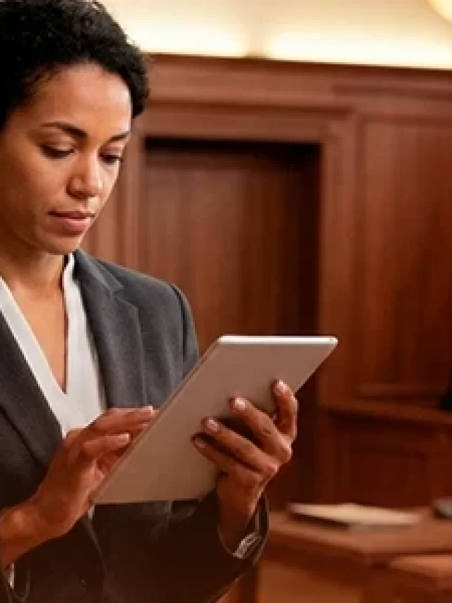 Mulher negra em tribunal usando tablet, representando profissão na área jurídica, com tribunal ao fundo e outros advogados presentes, demonstrando foco e profissionalismo.